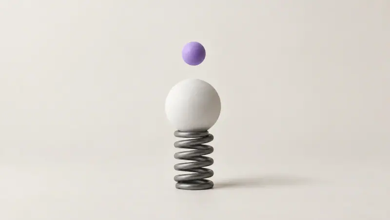 A white sphere rests on a compressed graphite coil spring while a smaller lavender sphere hangs in the air above it, mid-bounce.